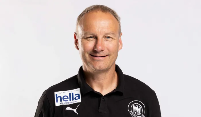 Ein Mann mit verschränkten Armen hinter dem Rücken der in die Kamera lächelt.