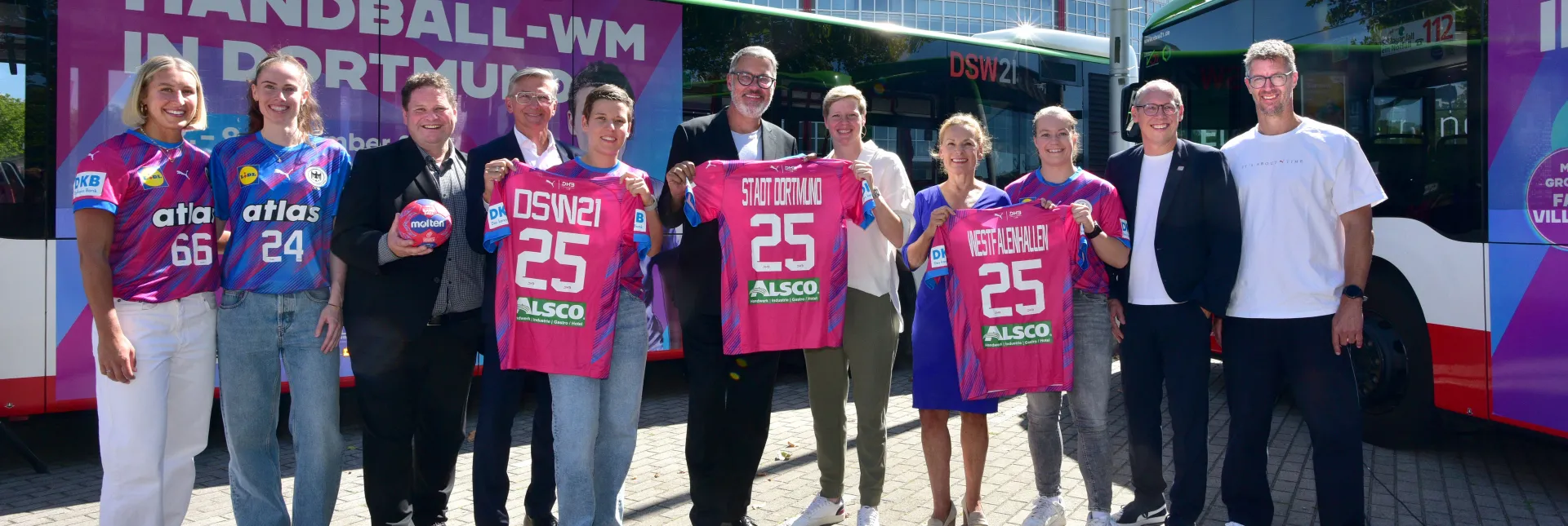 Spielerinnen und Verantwortliche von DHB, DSW21, Westfalenhallen und der Stadt Dortmund posieren mit Trikots und dem WM-Ball vor einem Bus im WM-Design