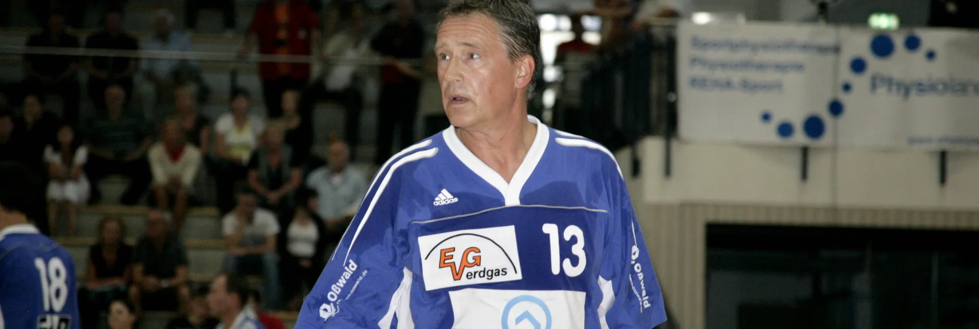 Ein älterer Mann in einem blauen Trikot der in einer Handballhalle steht.