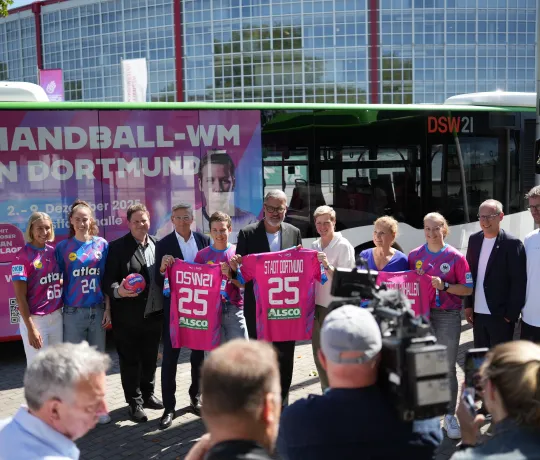 Gruppe posiert vor Westfalenhalle mit Bus im Handball-WM 25 Branding