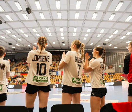 Eine Truppe von Handballerinnen in weißen Trikots die bei einem Tor aufstehen und jubeln