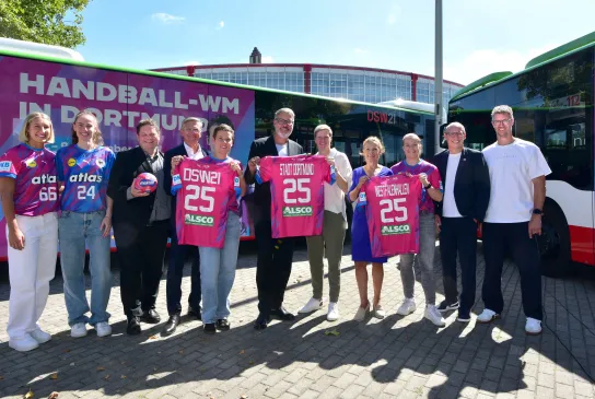 Spielerinnen und Verantwortliche von DHB, DSW21, Westfalenhallen und der Stadt Dortmund posieren mit Trikots und dem WM-Ball vor einem Bus im WM-Design