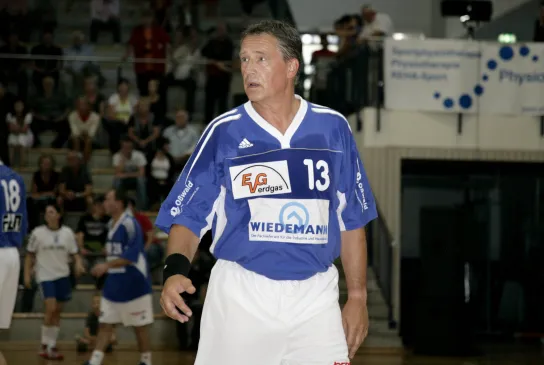 Ein älterer Mann in einem blauen Trikot der in einer Handballhalle steht.