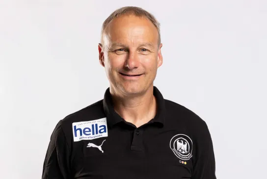 Ein Mann mit verschränkten Armen hinter dem Rücken der in die Kamera lächelt.
